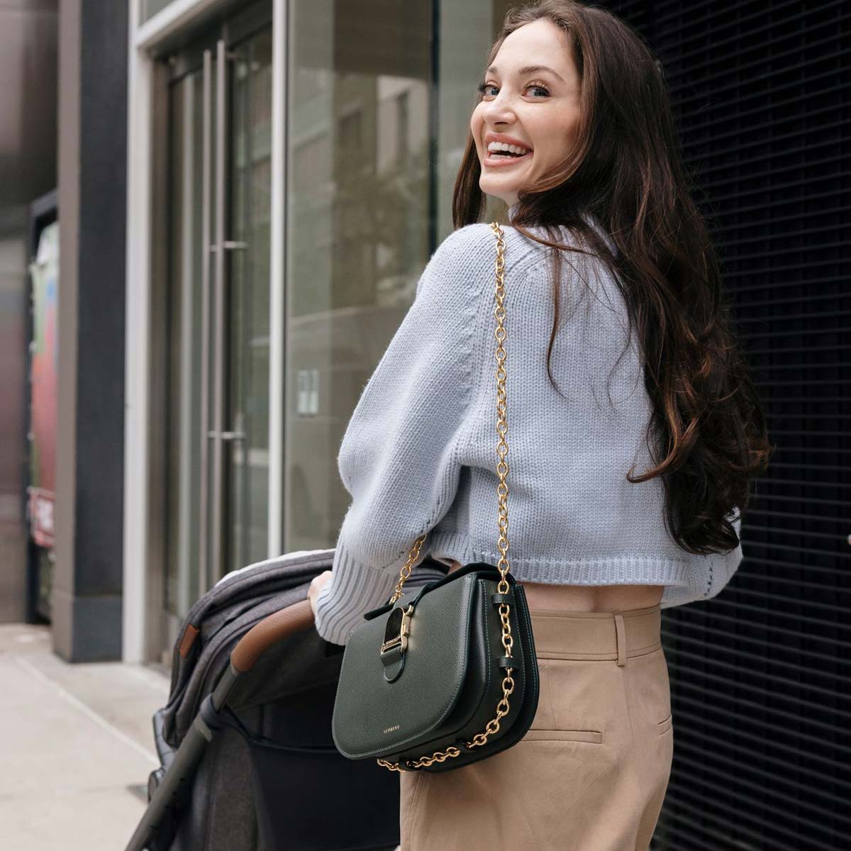 brunette mother pushing a stroller and wearing a green leather bag with a gold strap chain over her shoulder