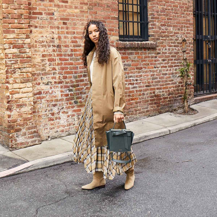 woman wearing a trench coat and plaid skirt holding a green bag by the top handle