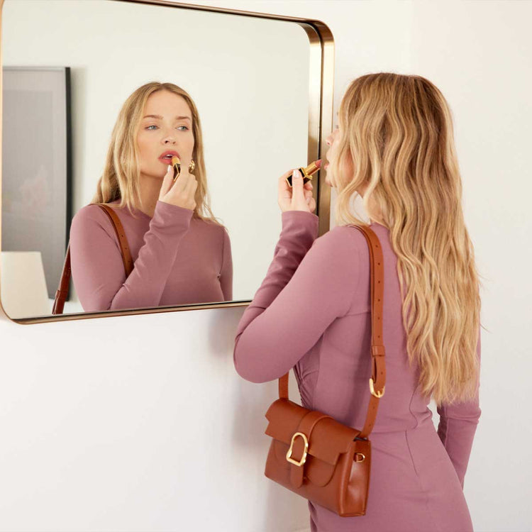 blonde woman wearing a long sleeve dress, putting on makeup with a brown leather belt bag over her shoulder