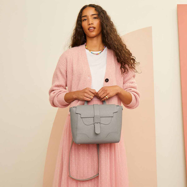 Model with light brown skin and dark wavy hair wears a pink cardigan and long tulle skirt, and holds a Midi Maestra Bag in Dolce Dove with Silver Hardware
