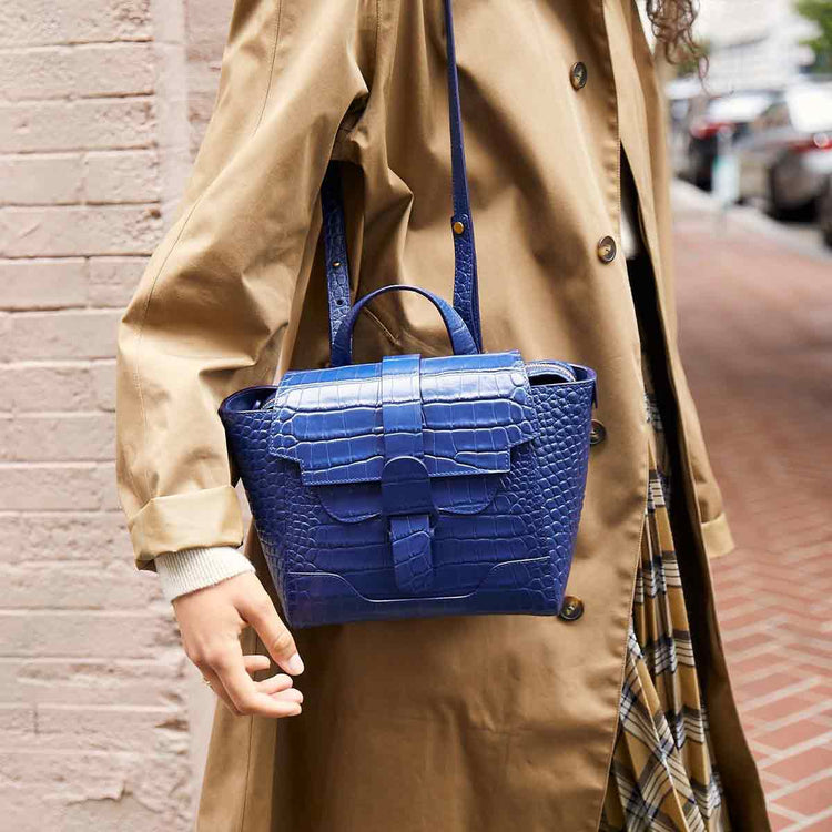 Mini Maestra Bag Marine with Gold Hardware Side, Shown on Shoulder of Woman with Pale Skin weaning a Tan Burberry Trench Coat 
