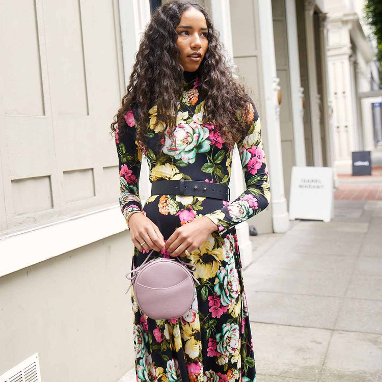 brunette model wearing a floral dress holding a purple leather bag by the top handle
