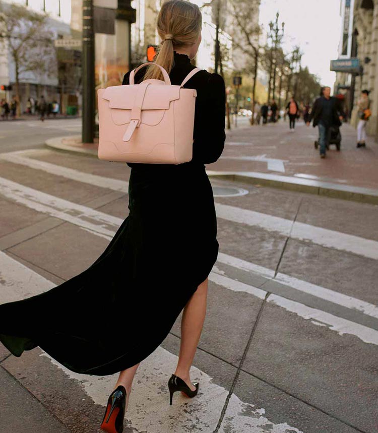 Woman wearing a long black dress and black heels with the Maestra Bag in Pebbled Blush as a backpack