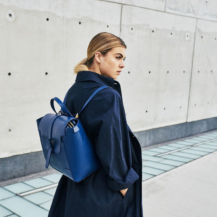 Blond woman in navy trench coat wears Maestra Bag Pebbled Marine with Gold Hardware as a backpack