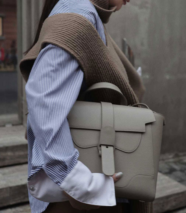 Woman wearing a blue button up shirt and brown knit vest and holding the Midi Maestra 