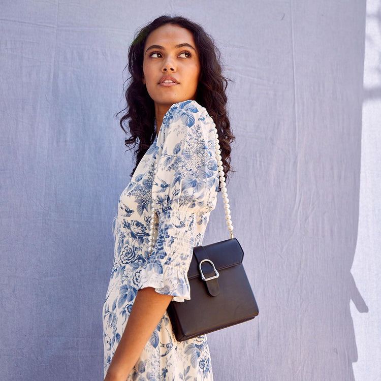 Pearl Shoulder Chain Attached to Cavalla Saddle Bag Piatta Noir on the Shoulder of Carmel Skinned Woman with Loose Curls and White and Blue Flower Patterned Dress