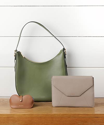 a large olive green hobo-style bag, a taupe leather tablet envelope sleeve, and a chestnut leather sunglasses holder sit on a wooden bench in front of a wall of white painted horizontal wood panels.
